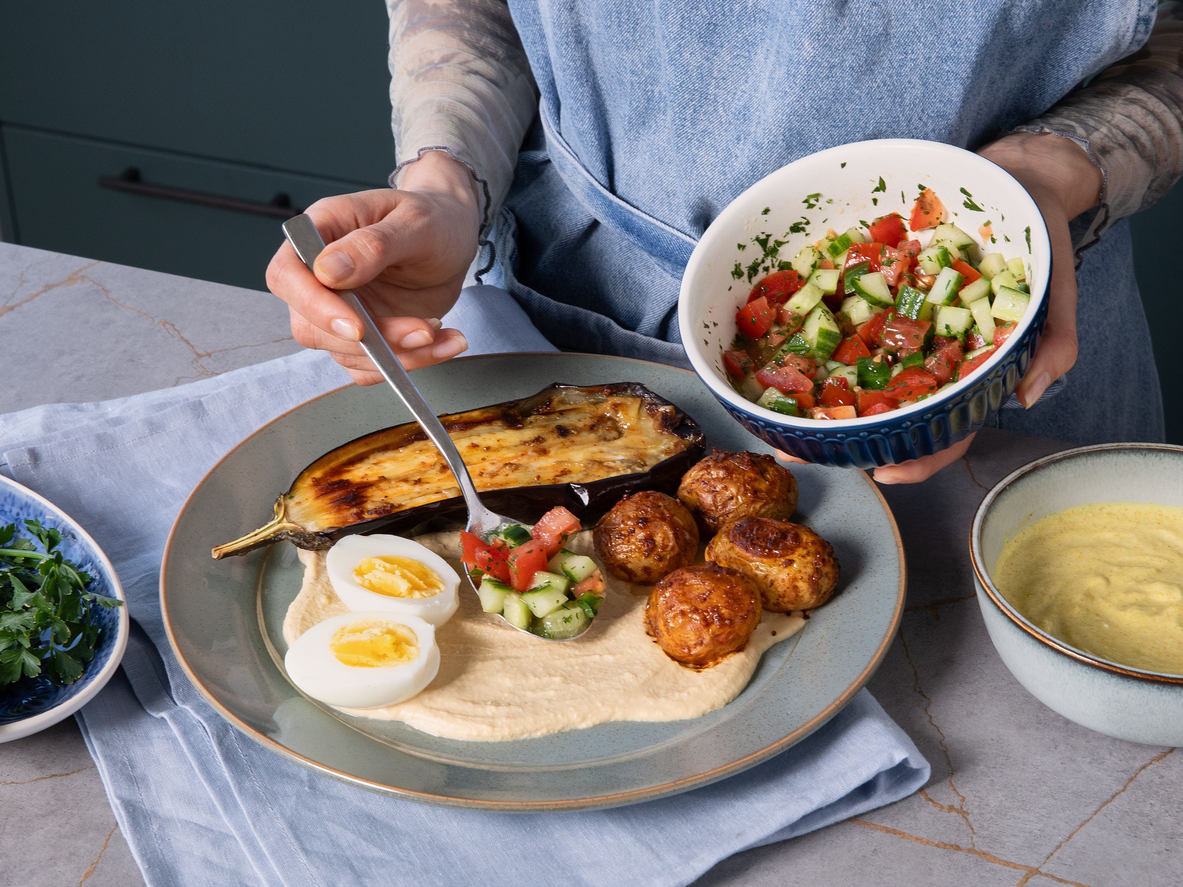 Den Hummus als Basis auf den Tellern verteilen. Auberginen, mit den Harissa-Kartoffeln, halbierten Eiern und dem Gurken-Tomaten-Salat darauf anrichten. Die Joghurtsauce darauf geben und optional noch mehr Harissa. Mit frischen Kräutern garnieren und mit warmem Fladenbrot servieren.
