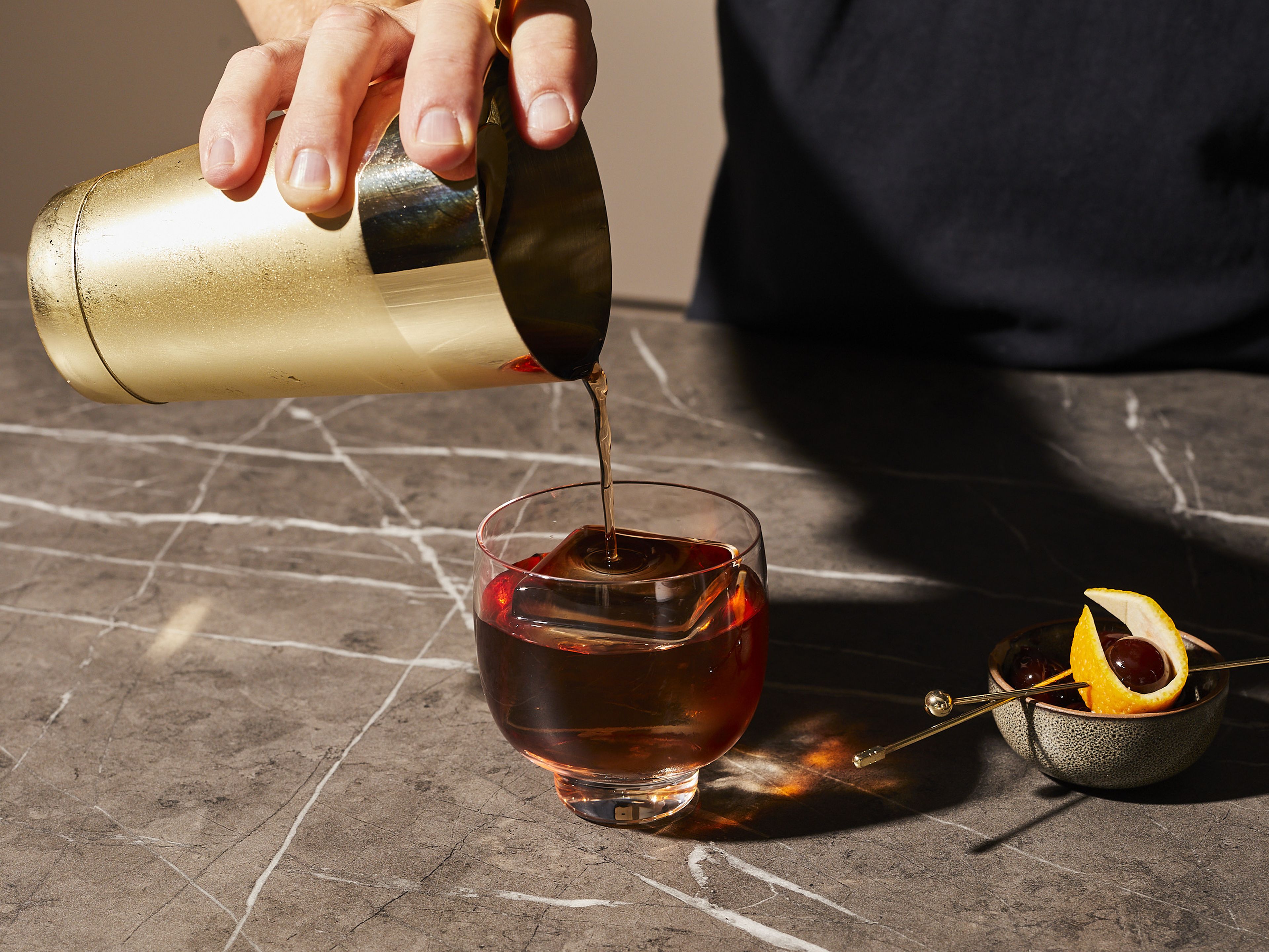 Einen großen Eiswürfel in das Glas geben. Das Cocktailsieb auf den Shaker legen, und den Drink in das Glas gießen. Mit einem Stückchen Orangenschale und einer Cocktailkirsche garnieren.