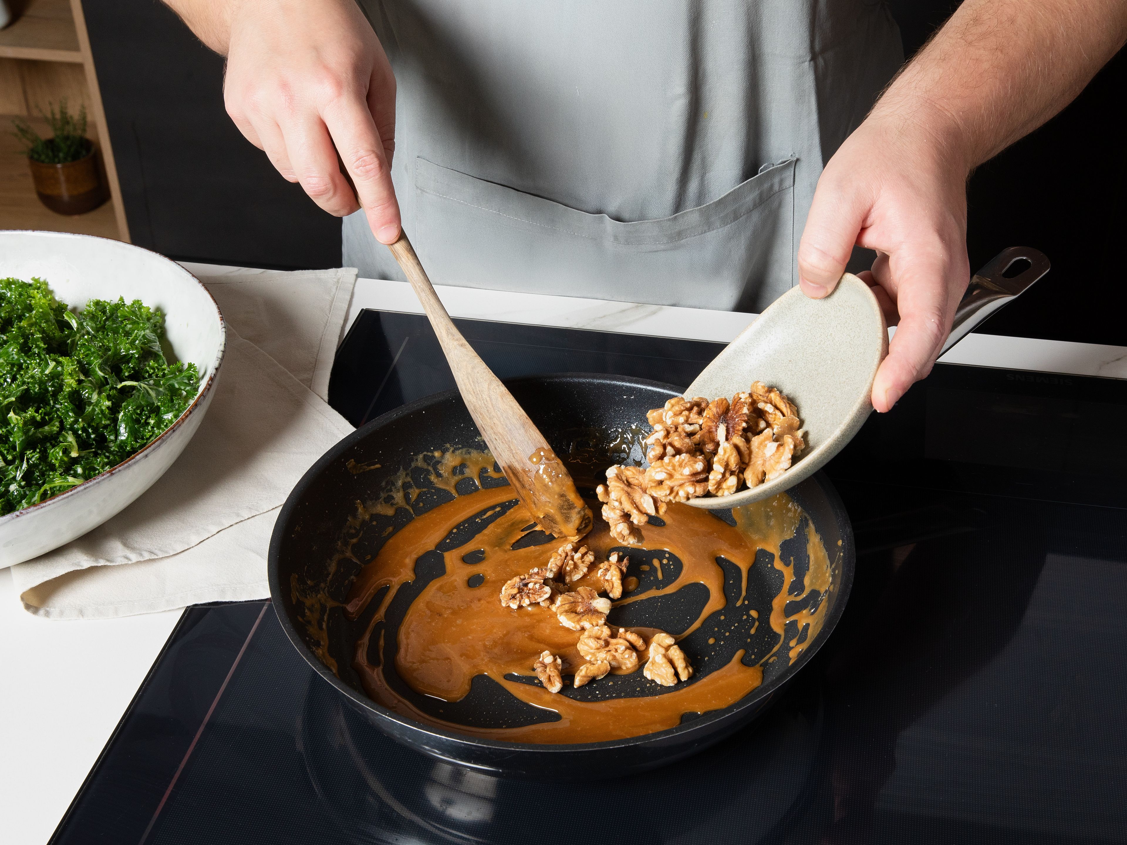 Die zuvor verwendete Pfanne auswischen. Die Pfanne auf niedrige-mittlere Hitze stellen, Zucker hinzugebe und schmelzen lassen. Butter dazugeben und auf mittlerer Hitze unter stetigem Rühren karamellisieren, bis die Masse eine hellbraune Farbe bekommt. Dann Walnüsse dazugeben und kurz unterrühren, bis sie komplett vom Karamell bedeckt sind. Auf Backpapier geben und vollständig auskühlen lassen, danach grob hacken. Apfel in dünne Scheiben schneiden. Äpfel, karamellisierte Walnüsse und Croutons zum Grünkohl geben. Zum Servieren noch etwas frischen Parmesan darüber hobeln.