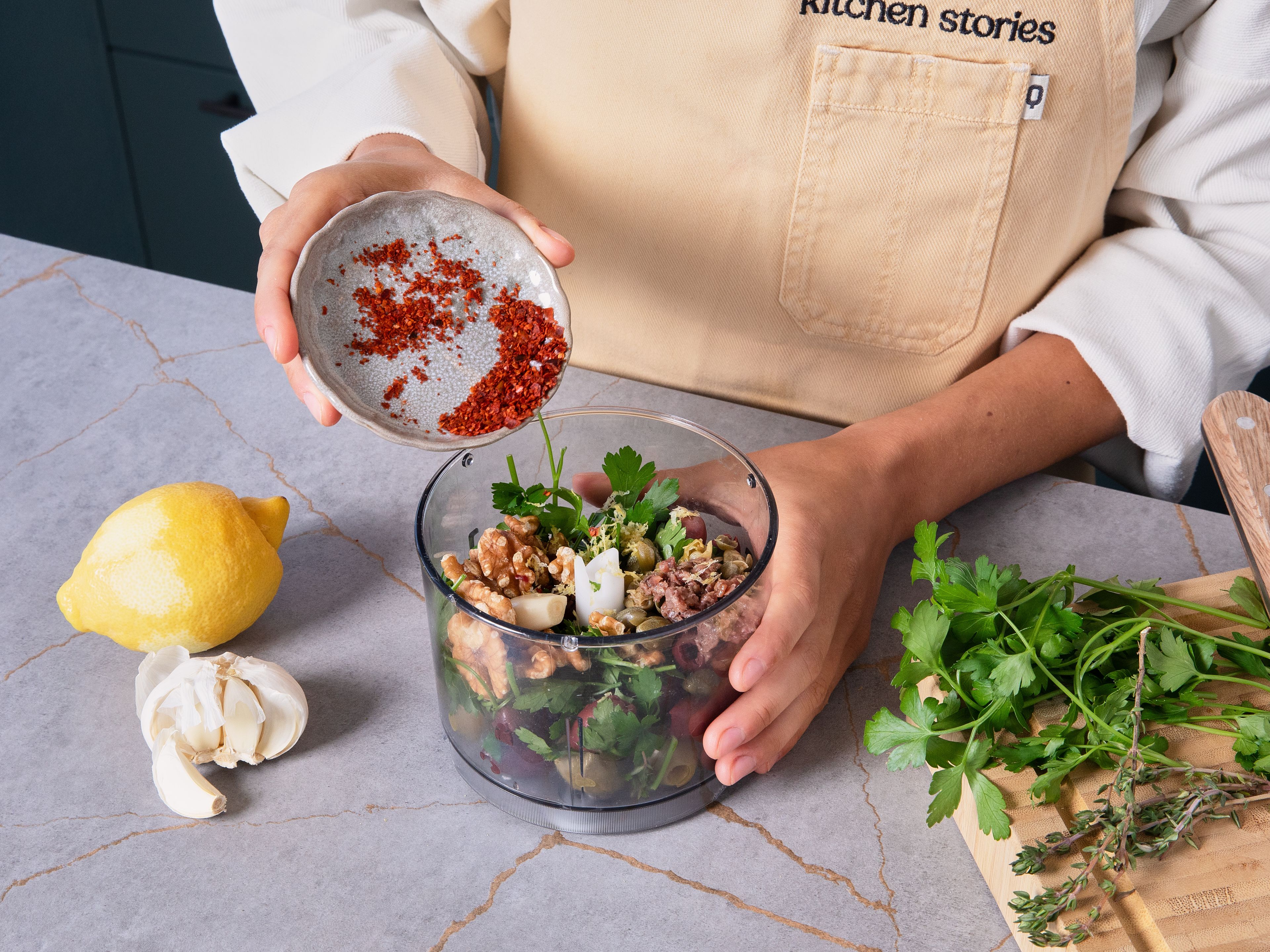 Die Blätter der Thymianstiele entfernen, die Zitrone schälen und auspressen. Knoblauch und Sardellen fein hacken und beides zusammen mit dem Zitronenabrieb, Petersilie, Thymian, Kapern, Oliven, Walnüssen und Chiliflocken in einen Zerkleinerer geben und kurz pulsieren, so dass es grob zerhackt ist, aber noch Struktur hat.