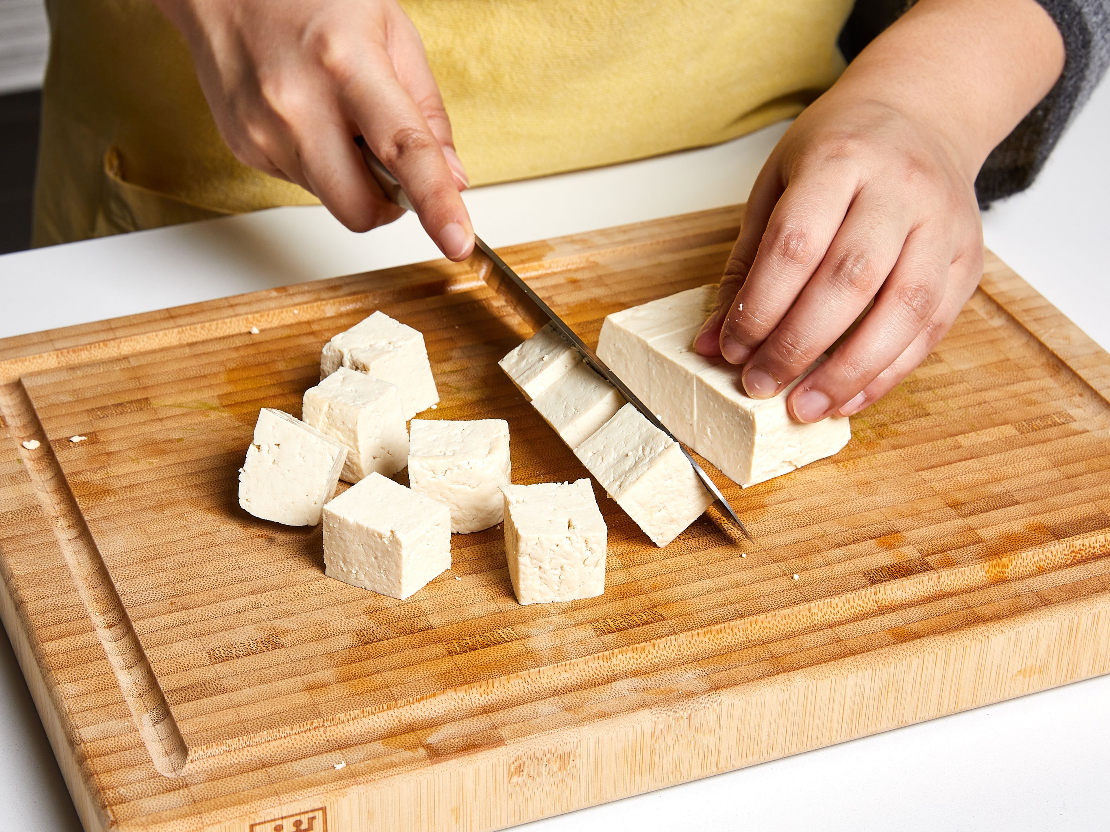 Tofu aus der Verpackung nehmen und abtropfen lassen. Den Tofu auf einem Teller mit Küchenpapier umwickeln. Einen schweren Gegenstand (eine Bratpfanne oder ein Backblech) darauf legen, um das Wasser herauszupressen. Ca. 20 Min. stehen lassen. In der Zwischenzeit grüne Bohnen von den Enden befreien und halbieren, Champignons vierteln. Schalotte in dünne Scheiben schneiden, Knoblauch und Ingwer fein hacken. Wenn der Tofu fertig ist, das Küchentuch entsorgen und den Tofu in ca. 2–3 cm große Würfel schneiden.