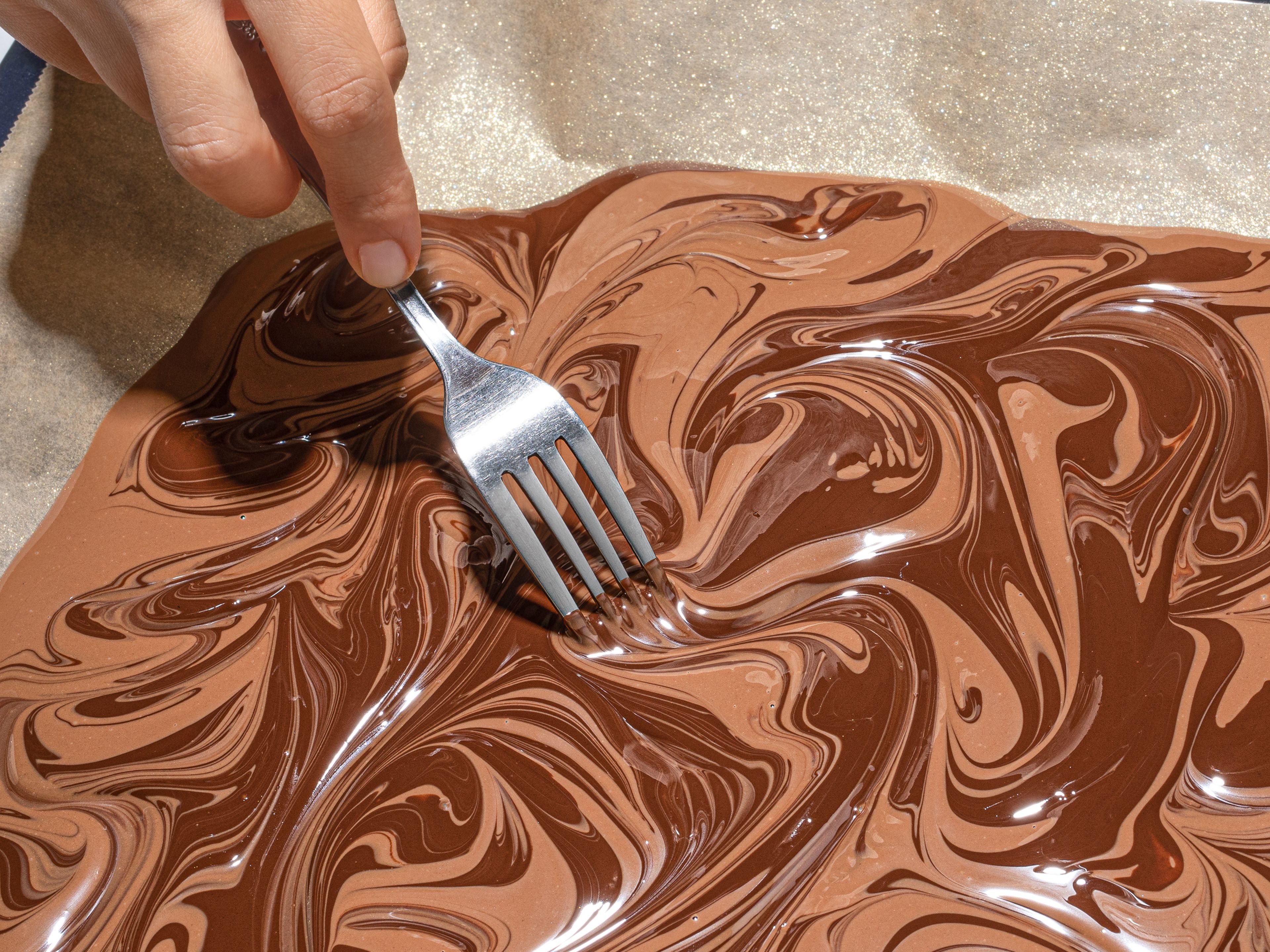 Die abgekühlte und geschmolzene Zartbitterschokolade auf dem Backpapier verteilen (ca. 0,25 cm dick) und anschließend die abgekühlte und geschmolzene Vollmilchschokolade gleichmäßig darauf verteilen. Mit einer Gabel in kreisenden Bewegungen die beiden Schokoladen miteinander verbinden, um einen Marmoreffekt zu kreieren. Um kleine Luftbläschen in der fertigen Bruchschokolade zu vermeiden, das Blech mehrmals vorsichtig auf die Arbeitsplatte klopfen. Für ca. 5 Min. ruhen lassen.