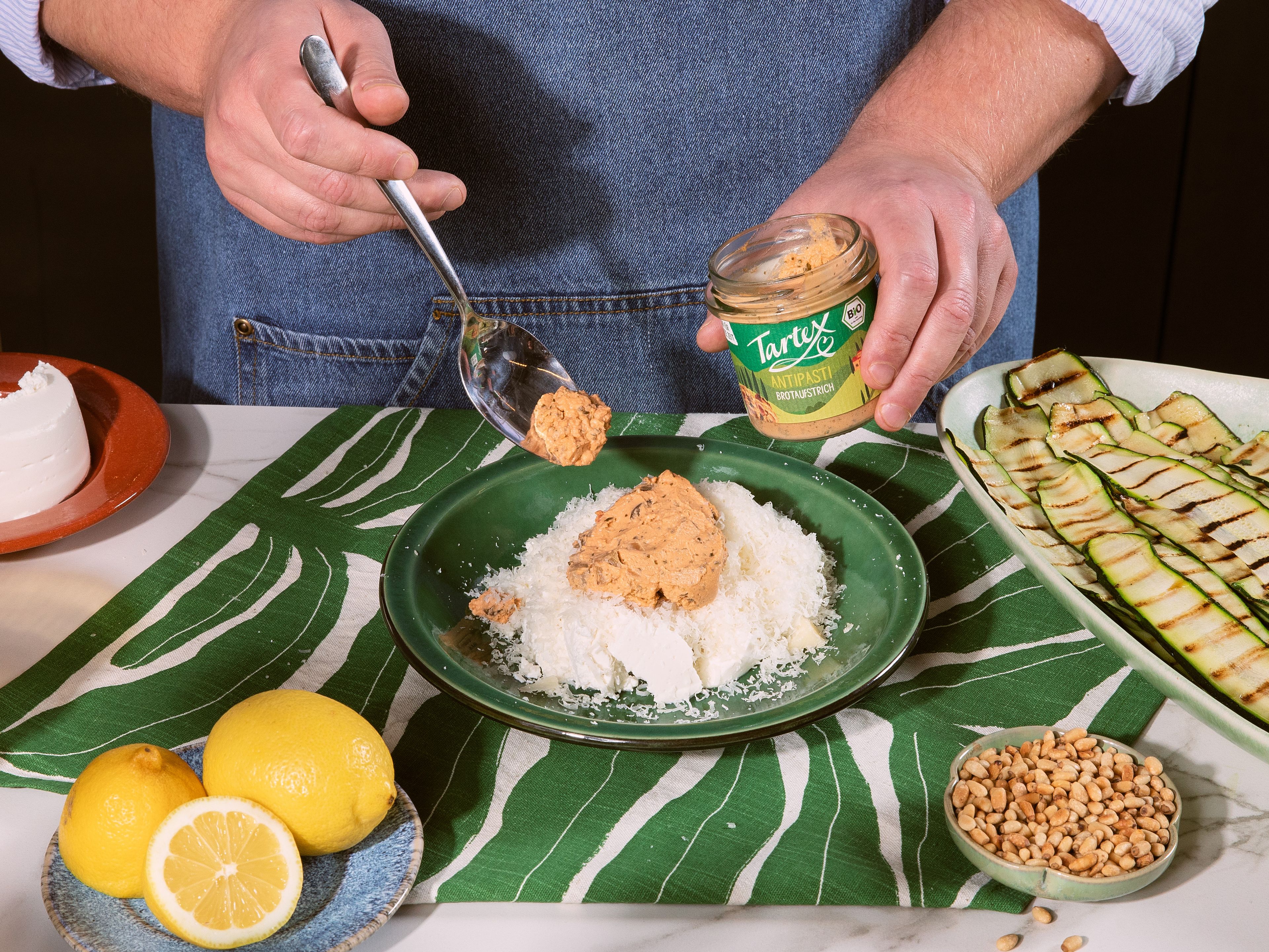 Basilikum grob hacken und Parmesan fein reiben. In einer Schüssel Tartex Antipasti Brotaufstrich, Ricotta, Parmesan, die Hälfte der Pinienkerne, Zitronenabrieb, ca. 4 EL Zitronensaft und die Hälfte des Basilikums vermengen. Jeweils ca. 1 EL der Masse auf das Ende eines Zucchinistreifens geben und aufrollen.