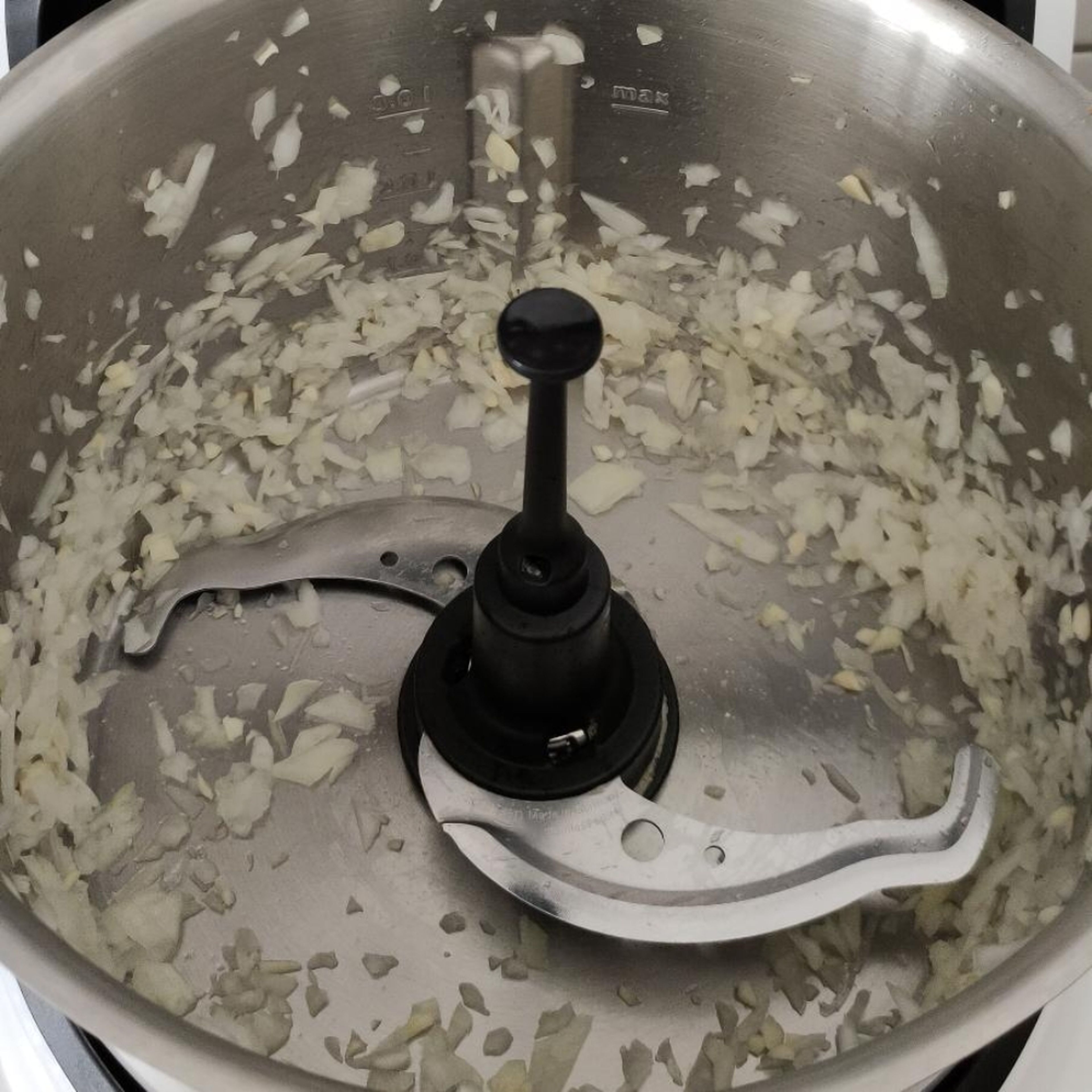 Universalmesser in den Cookit einsetzen. Zwiebel und Knoblauch schälen und halbiert in den Topf geben. Deckel schließen, Messbecher einsetzen und klein schneiden (Universalmesser | Stufe14 | 10 Sek.).