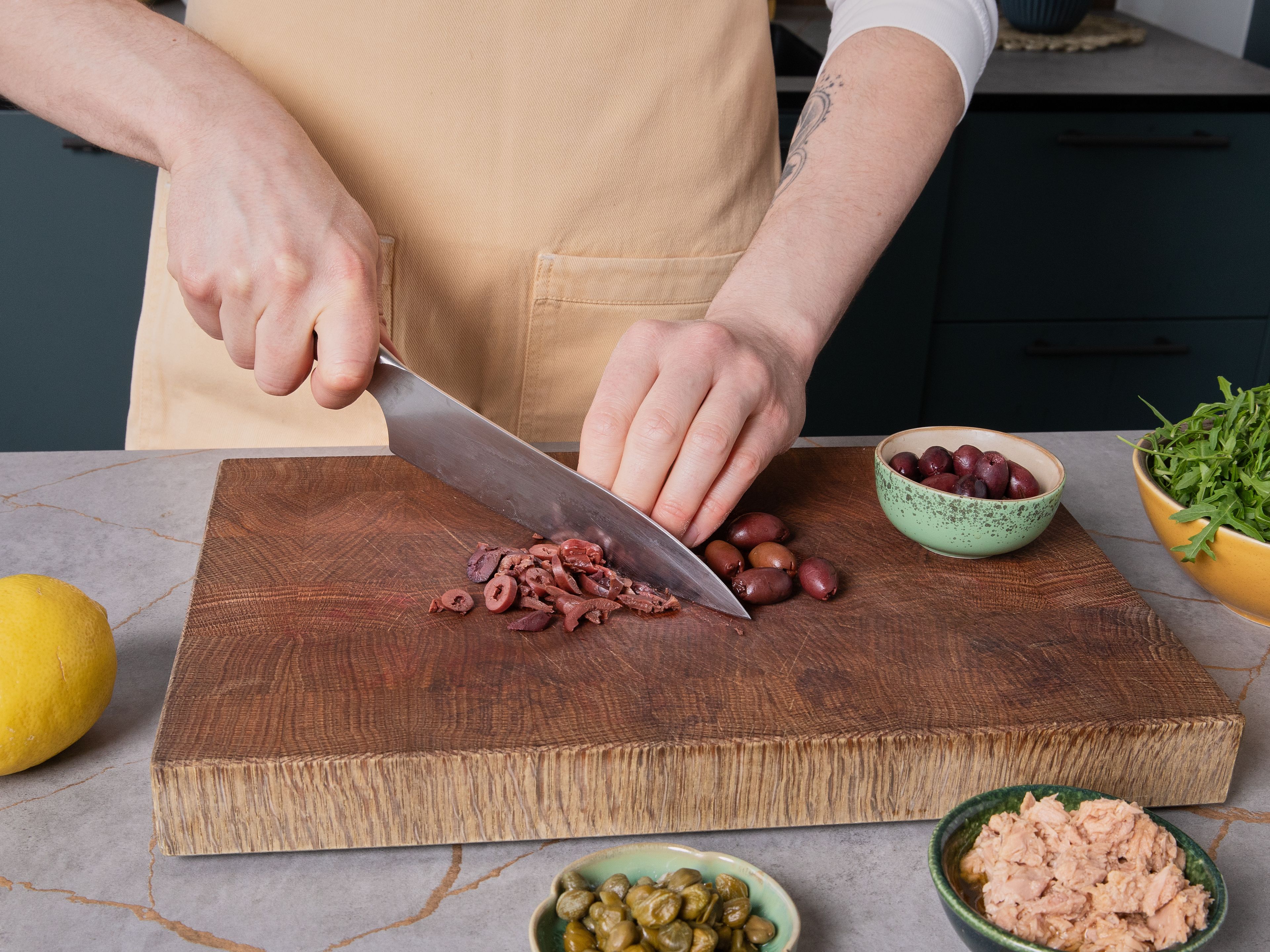 Einen großen Topf mit gesalzenem Wasser zum Kochen bringen und die Pasta darin nach Packungsanweisung al dente kochen. Währenddessen Oliven entsteinen und grob hacken, Kapern abtropfen lassen und ebenfalls grob hacken. Den Rucola waschen, trocknen und grob schneiden. Parmesan fein reiben. Die Zitrone heiß abwaschen, etwas Schale abreiben und den Saft auspressen.