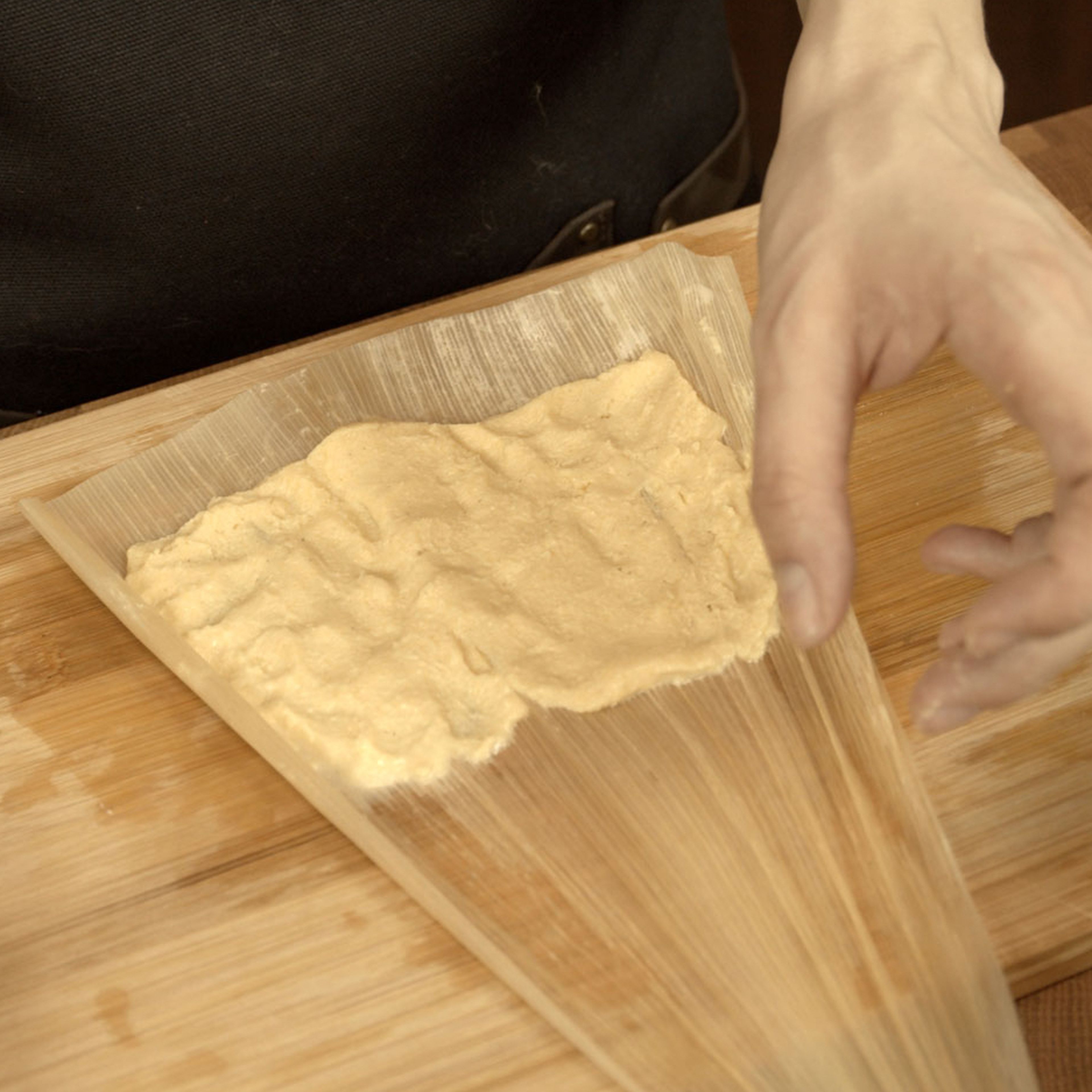 Start assembling the tamales. Lay corn a soaked corn husk glossy side up. Add 2 tbsp of masa dough in the wider half of the corn husk. Spread it evenly in the wider half, leave 1-2 cm gap from top and sides. Tip: dip your fingers in a bowl with clean water, it will prevent dough from sticking