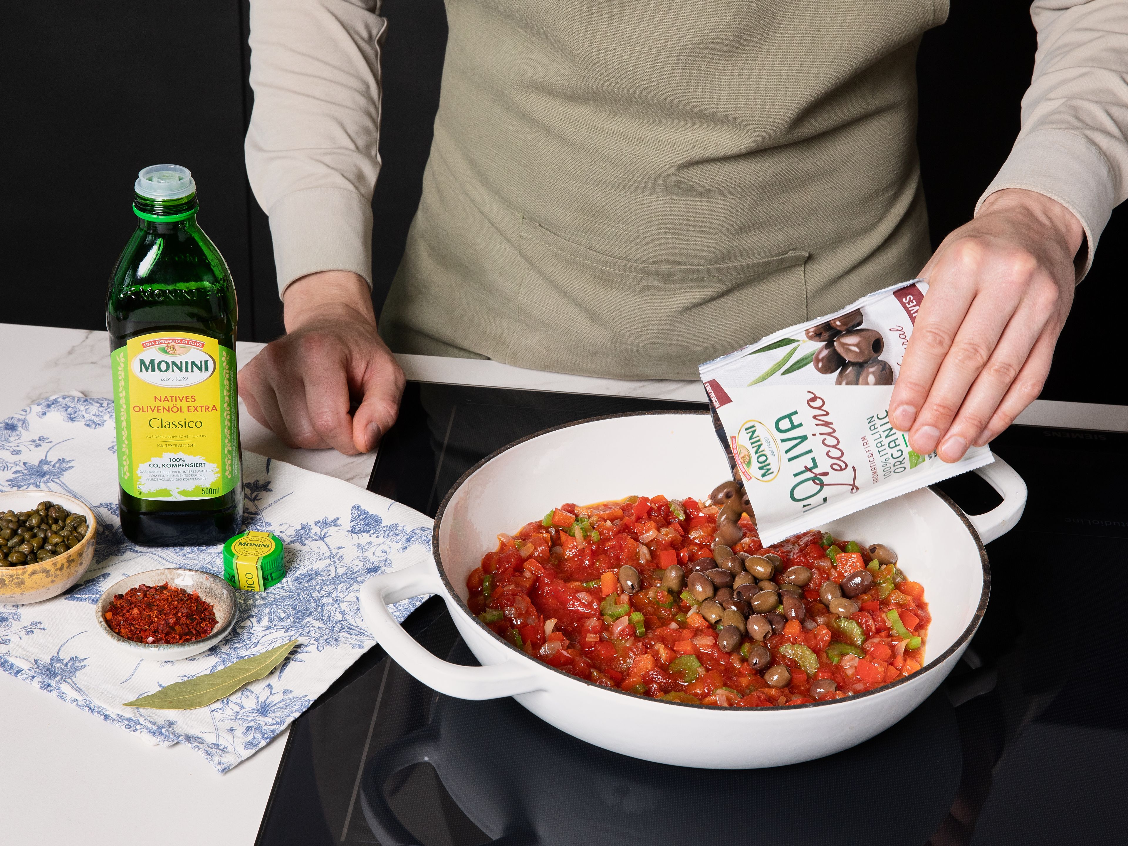 In einer großen Pfanne das restliche Monini Classico Olivenöl erhitzen. Zwiebeln, Sellerie und Paprika anbraten, bis sie weich sind. Dann Tomaten, Monini Schwarze Oliven, Kapern, Rosinen, Lorbeerblatt und Chiliflocken hinzufügen. Mit Salz und Pfeffer würzen und 10 Minuten köcheln lassen. Anschließend Rotweinessig und Weißwein dazugeben und weitere 5 Minuten köcheln lassen.