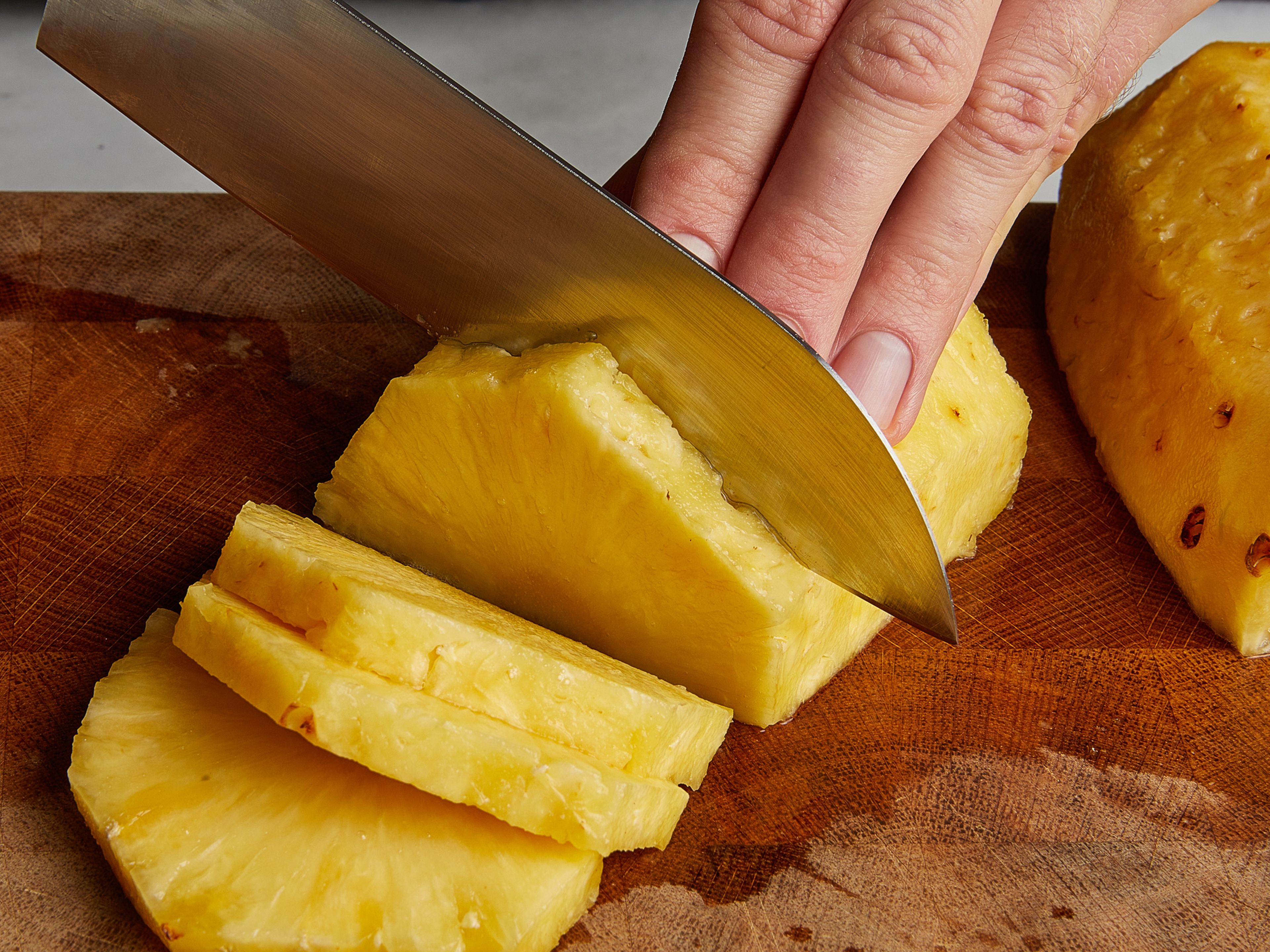 Beide Enden der Ananas abschneiden. Ananas hochkant stellen und die Schale ringsherum großzügig abschneiden. Die Ananas halbieren, in Scheiben schneiden und den Strunk aus der Mitte herausschneiden.