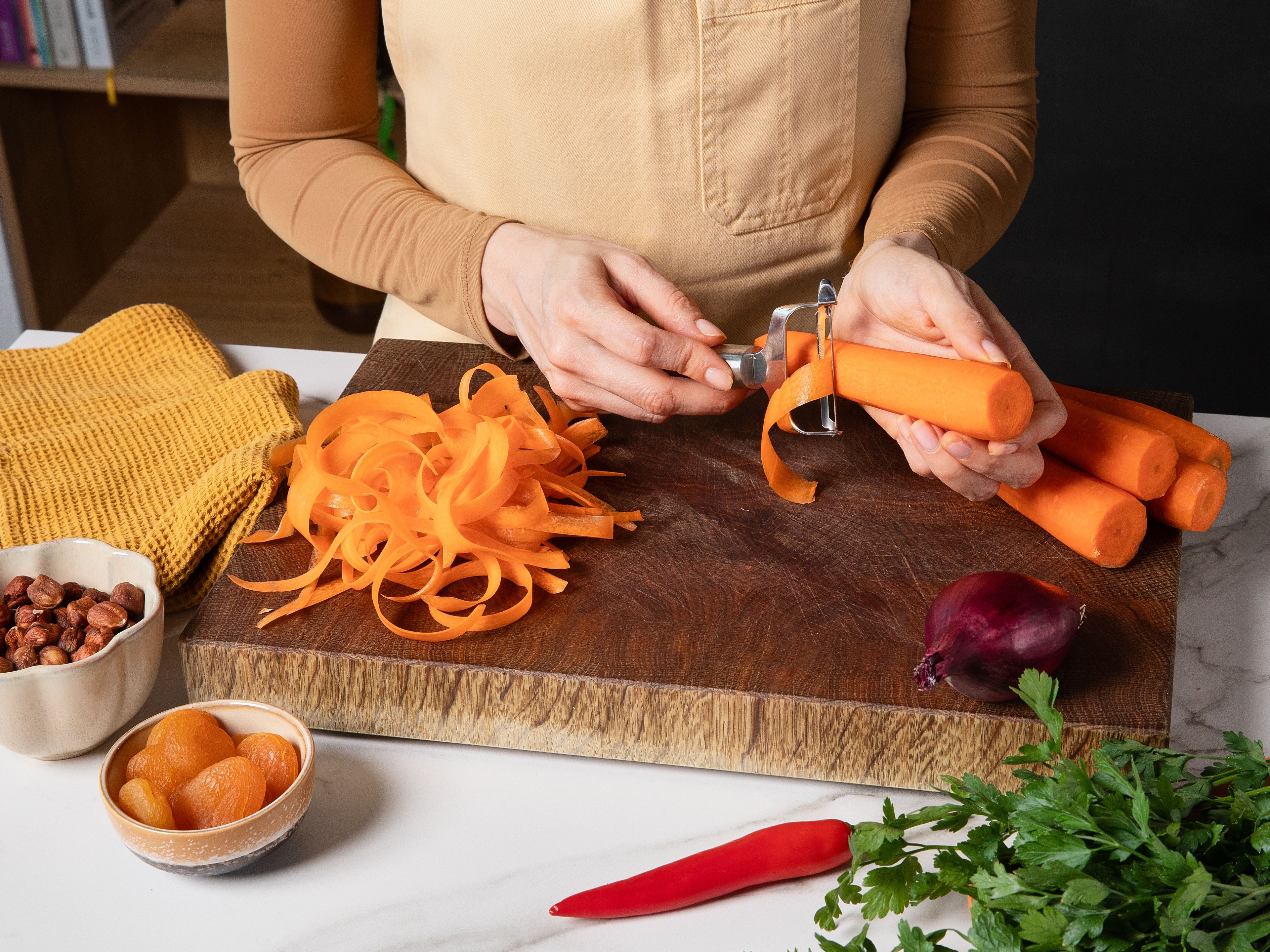 Währenddessen die Karotten schälen und dann mit einem Sparschäler in feine Streifen schneiden. Rote Zwiebel halbieren und in feine Ringe schneiden. Getrocknete Aprikosen in feine Streifen schneiden und alles in eine Servierschüssel geben. Haselnüsse und Petersilie grob hacken und beiseite stellen. Optional Chili in feine, schräge Ringe schneiden, falls verwendet.