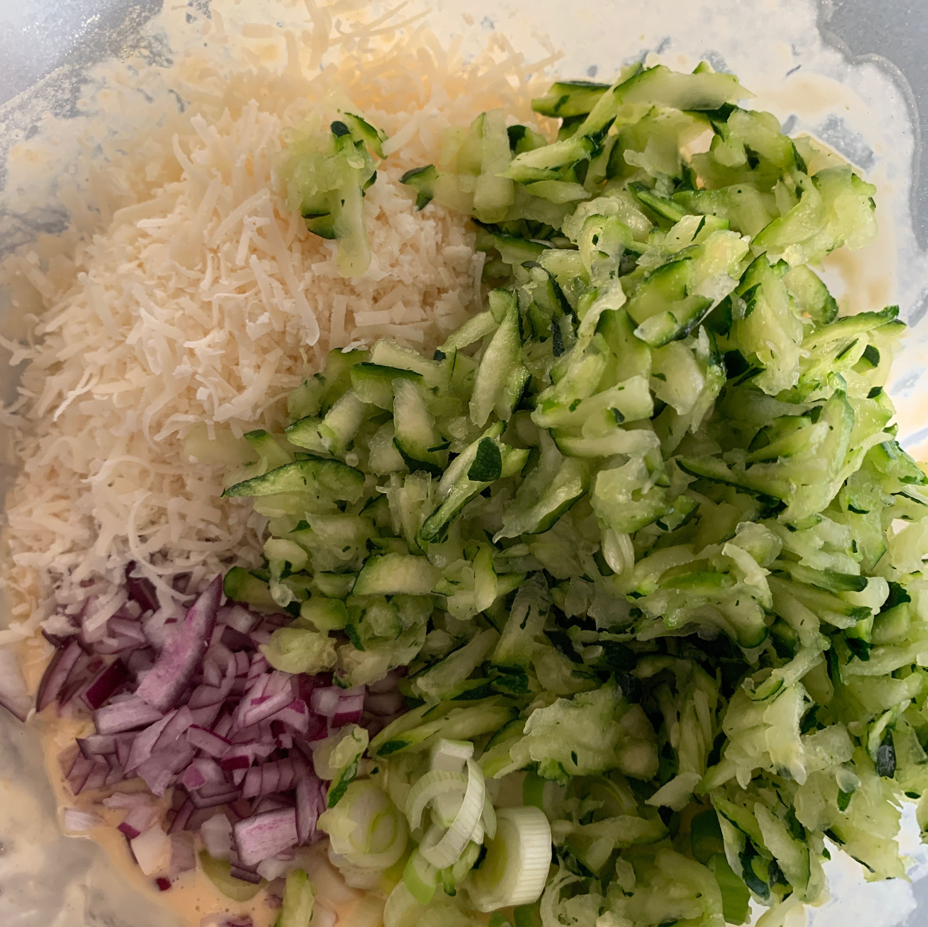 Zucchini, Parmesan, Frühlingszwiebel, Knoblauch und rote Zwiebel in den Teig geben. Mit Salz und Pfeffer würzen und vermischen.