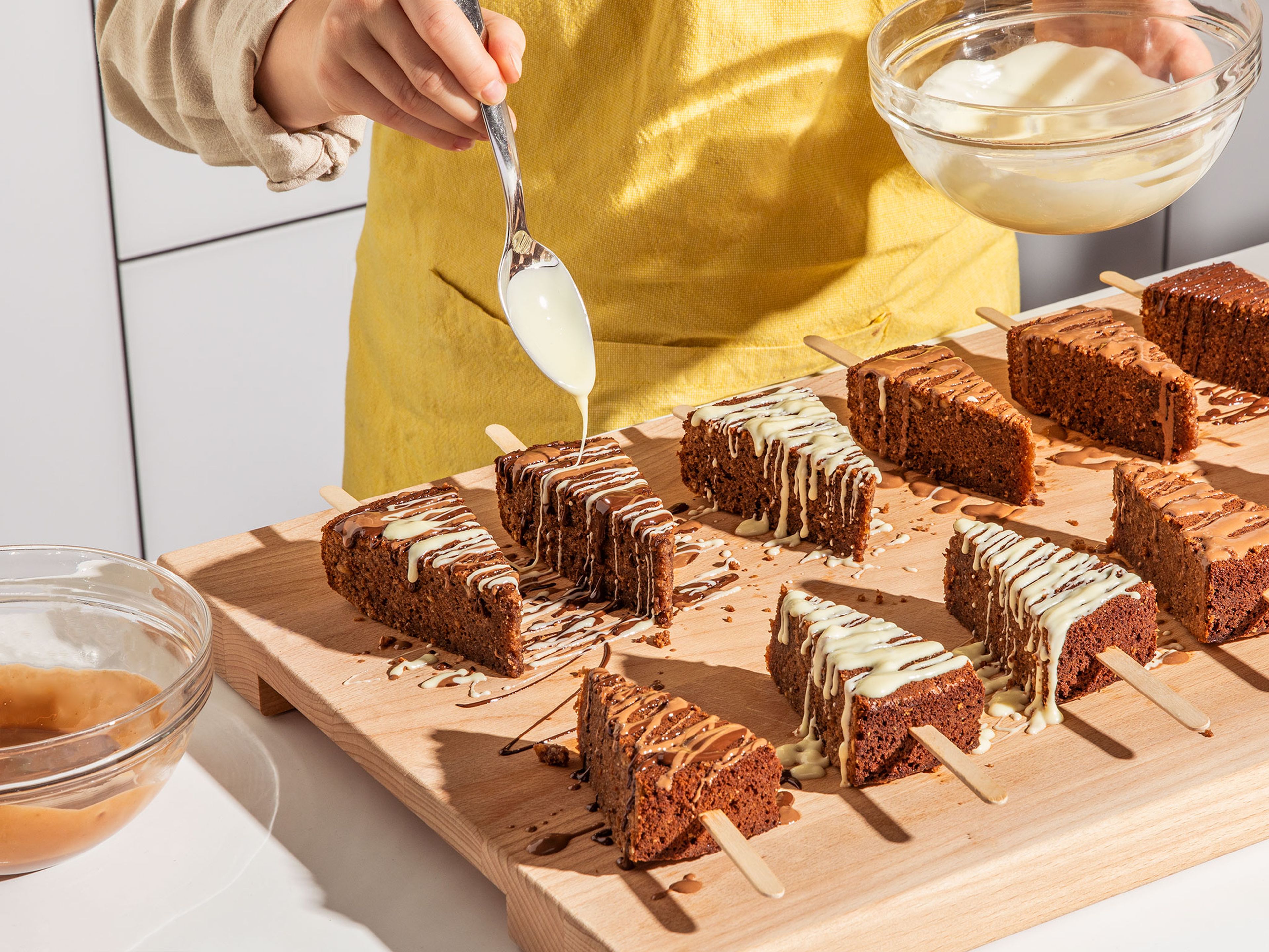 Den abgekühlten Kuchen in 12 gleich große Stücke schneiden und jeweils ⅓ eines Eisstiels in das Kuchenstück schieben. Mit allen Stücken wiederholen. Die Kuchen am Stiel nach Belieben mit den verschiedenen Schokoladen, gehackten Haselnüssen und z.B. Ferrero Küsschen oder getrockneten Blüten dekorieren. Der Kreativität sind keine Grenzen gesetzt. Anschließend für ca. 30 Min. in den Kühlschrank stellen, damit die Schokolade fest wird.