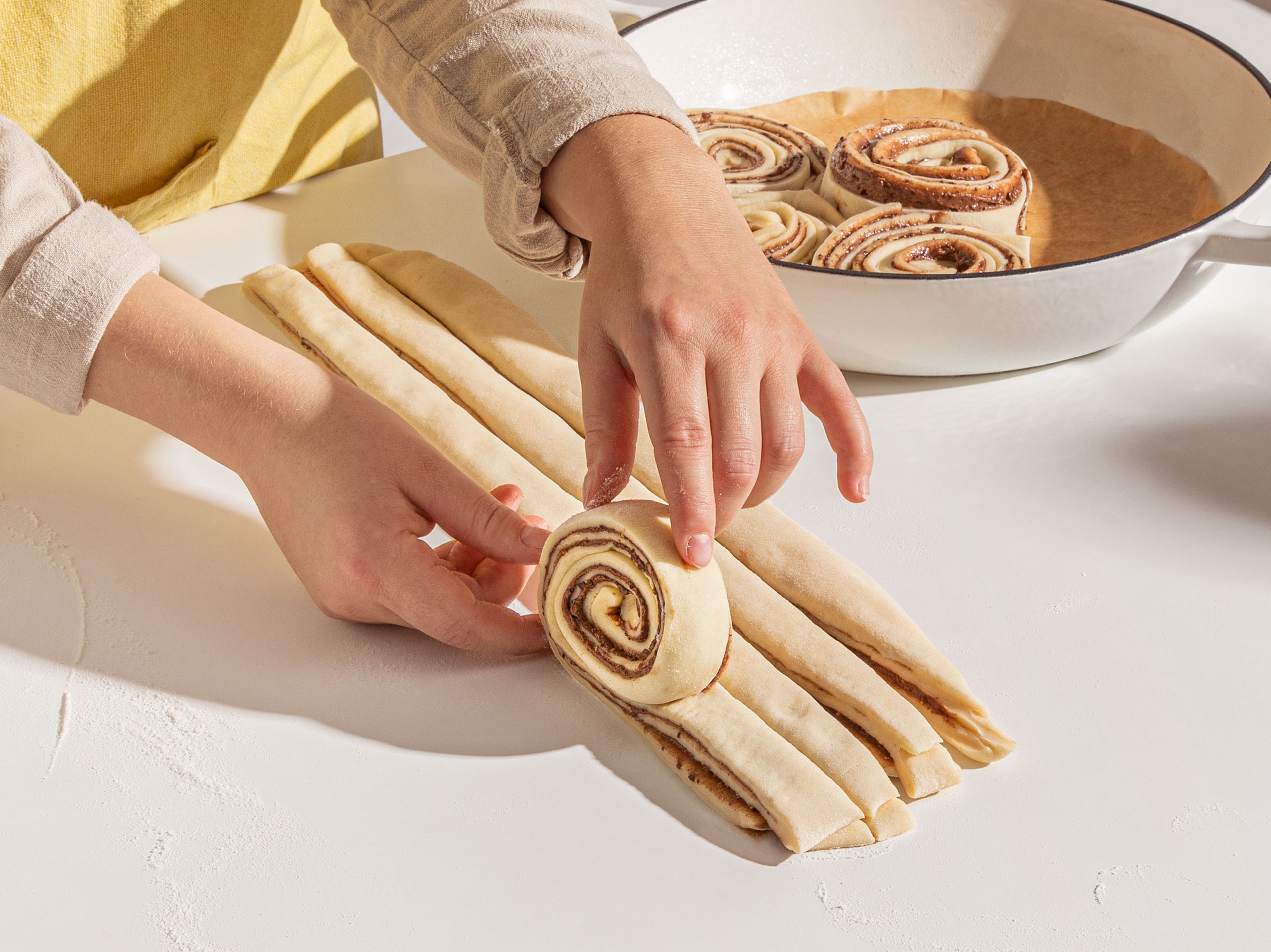 Nach der Ruhezeit wird der Teig mit Hilfe des Nudelholzes noch etwas flacher ausgerollt. Mit einem Messer oder Pizzaschneider kann das Rechteck nun längs in 8 gleich dicke Stränge geschnitten werden. Die Stränge werden anschließend einzeln aufgerollt, und das Ende der Schnecke auf der Unterseite versteckt. Die fertigen Schnecken werden kreisförmig in eine mit Backpapier ausgelegte runde Backform platziert und ruhen locker abgedeckt mit einem Geschirrtuch, für ca. 1 Std. Nach dem Ruhen sollten sich die Schnecken berühren.