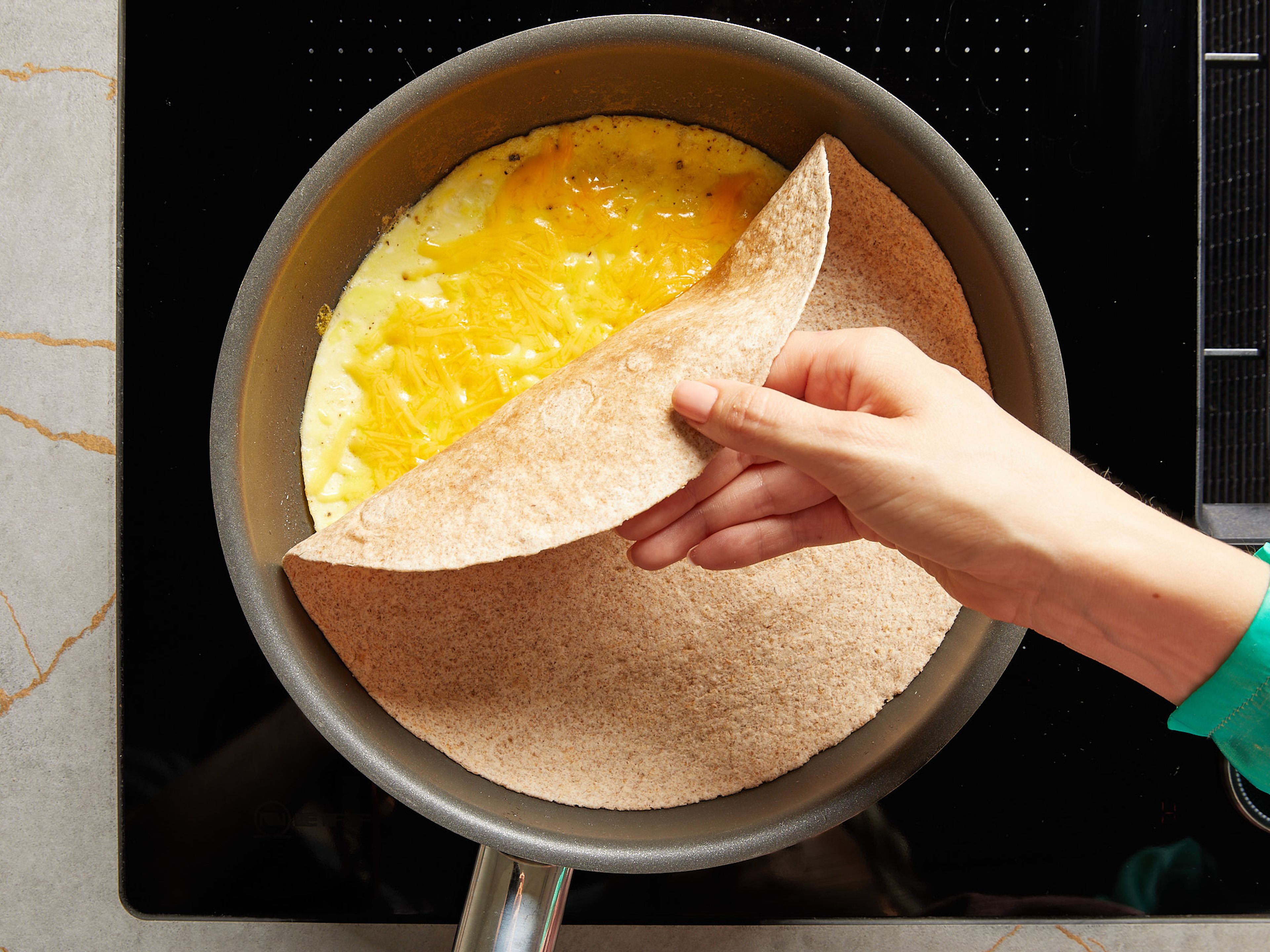 Sobald das Ei stockt, aber oben noch leicht flüssig ist, den geriebenen Käse darauf verteilen. Wenn der Käse etwas angeschmolzen ist, die Tortilla darauf legen. Und bei geschlossenem Deckel ca. 1–2 Min. weiter braten lassen, damit das restliche Ei stockt und die Tortilla am Käse haften bleibt. Dann aus der Pfanne auf die Arbeitsplatte stürzen, sodass die Ei-Schicht oben liegt.