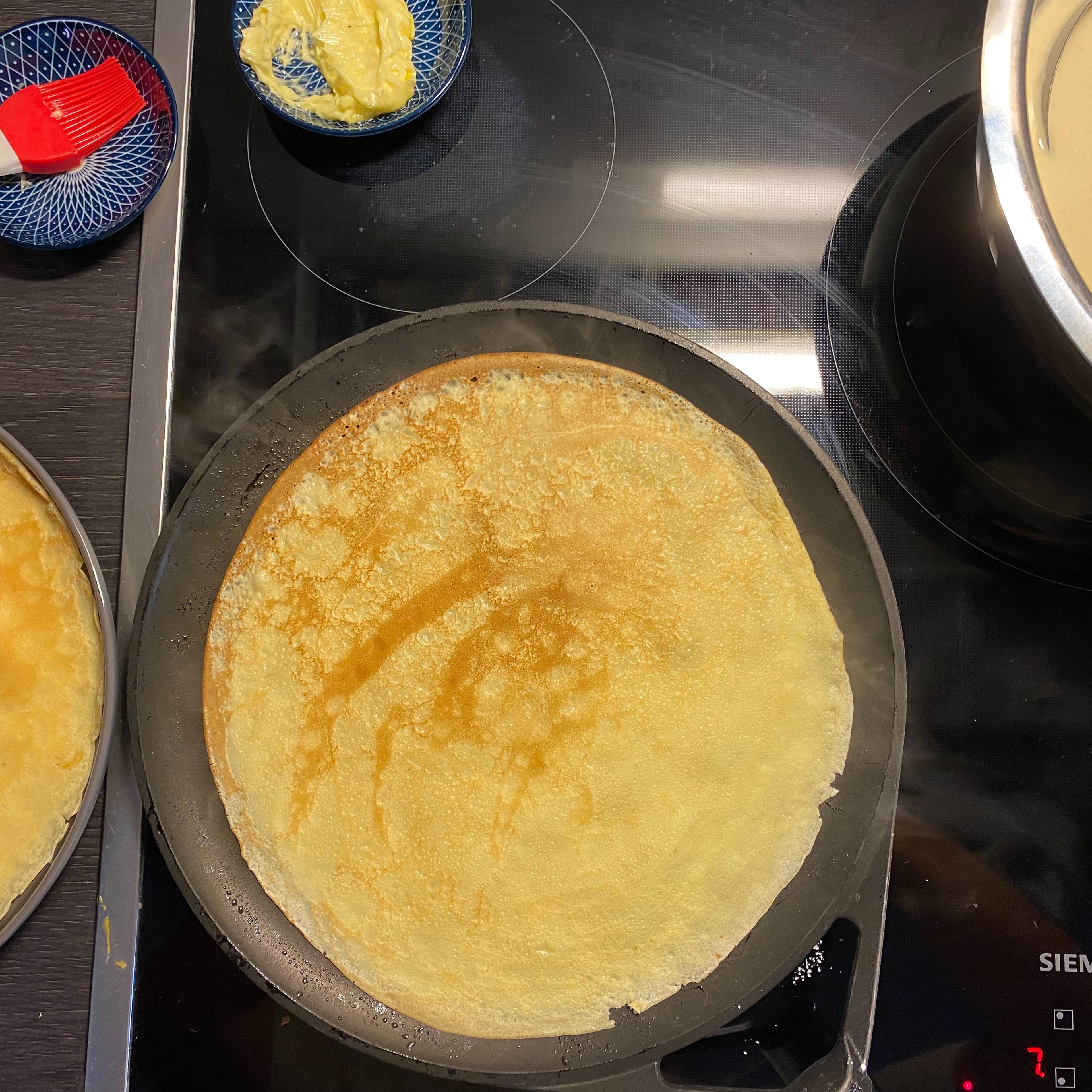 Nachdem der Teig geruht hat können die Eierkuchen bzw. Pfannkuchen ausgebacken werden. Bei jedem zweiten wird zunächst die vorgeheizte Pfanne (mittlere bis hohe Hitze) mit Butter bestrichen. Nun kommt eine Kelle Teig in die Pfanne, welche dann kreisförmig geschwenkt wird, damit die Eierkuchen schön dünn werden. Jede Seite für etwa 1 min goldbraun braten und dann auf einem Teller zur Seite stellen.