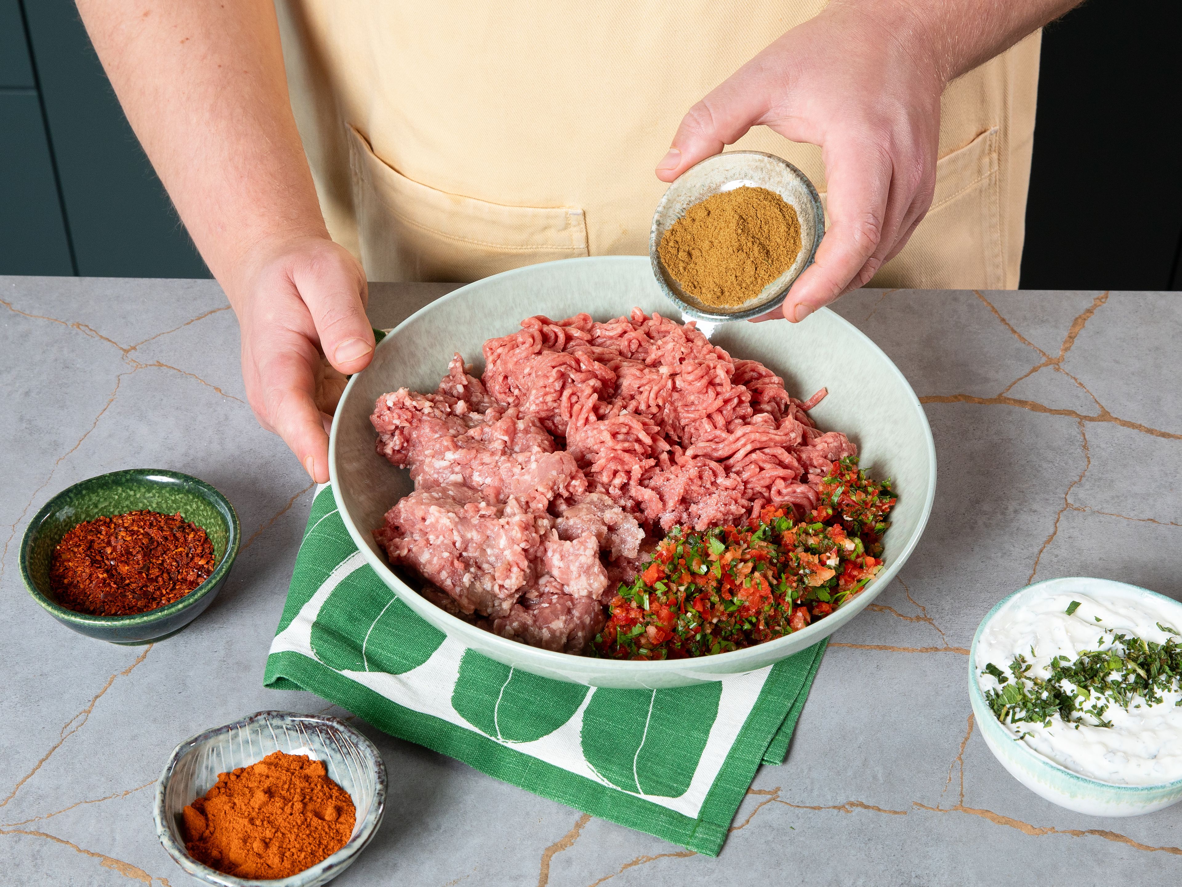 Das Hackfleisch dazugeben und mit Paprikapulver, Kreuzkümmel, Chiliflocken und Salz würzen. Gut durchkneten und für ca. 1 Std. in den Kühlschrank stellen. Dann jeweils eine Handvoll von der Hackmasse an einen Spieß drücken. Vor allem die Enden sollten fest angedrückt werden.